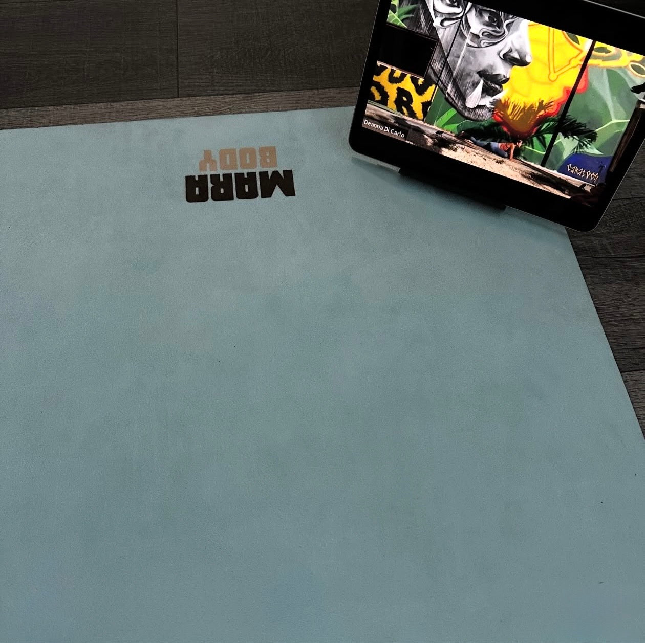 Vegan suede aesthetic blue yoga mat with the brand name 'MARA BODY' shown on a dark wood floor next to a tablet displaying a yoga poses fitness app