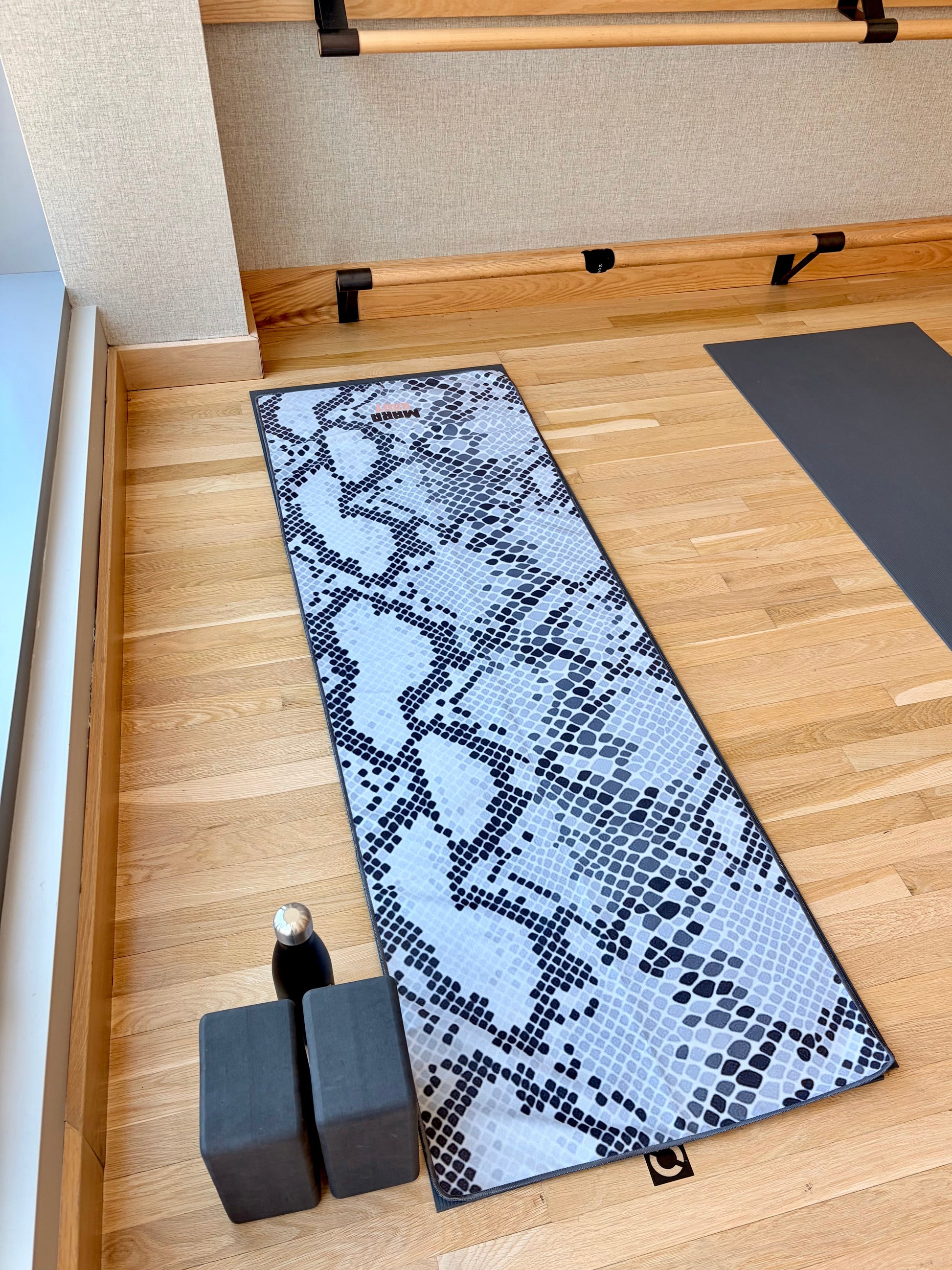 A yoga towel over a yoga mat with a black and white mosaic snake skin pattern is laid out on a light wood floor in a hot yoga studio setting, alongside two dark gray yoga blocks and a small bottle.