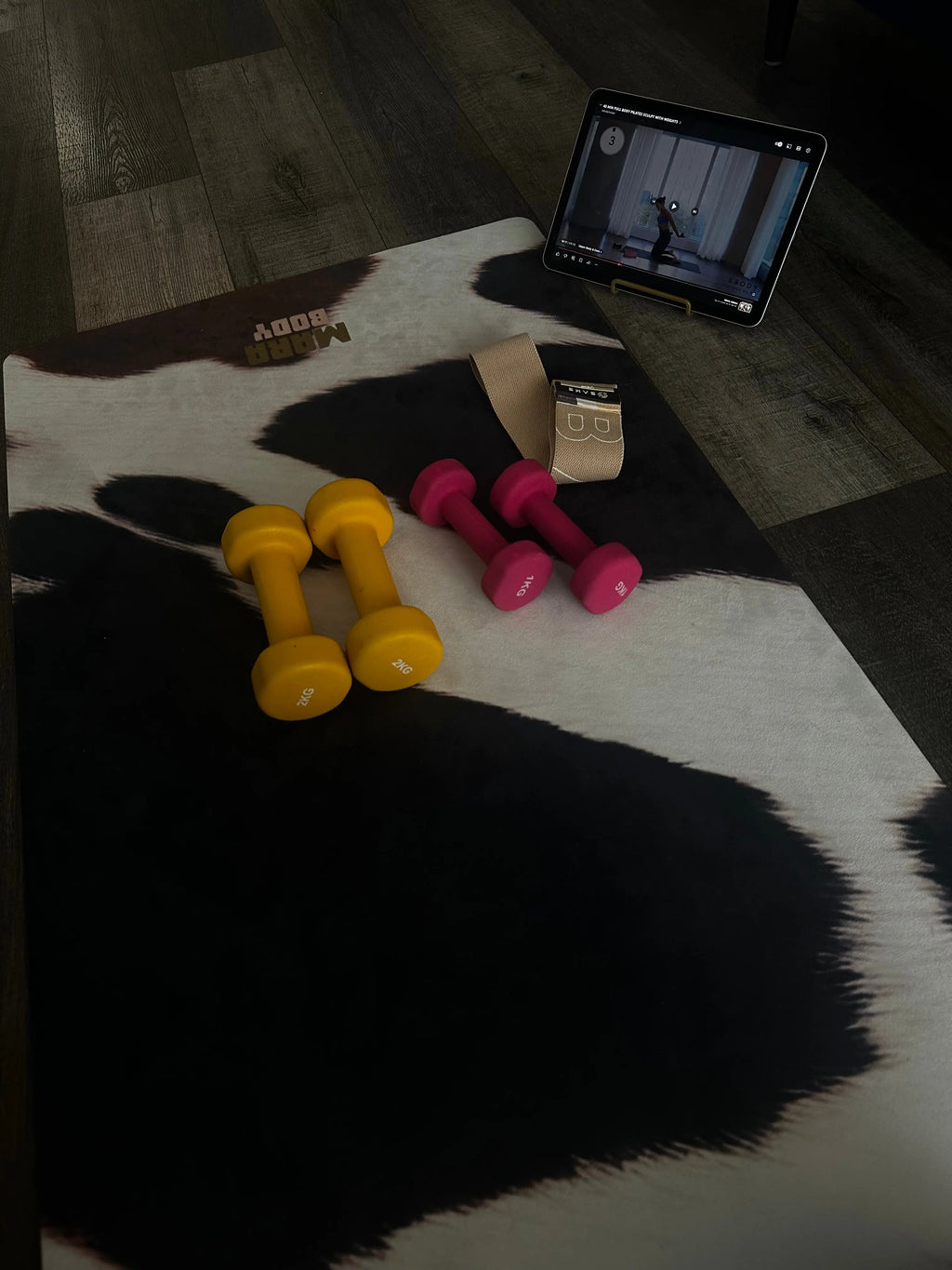 Two yellow and two pink dumbbells on a cowhide yog amat with a tablet showing a fitness app.