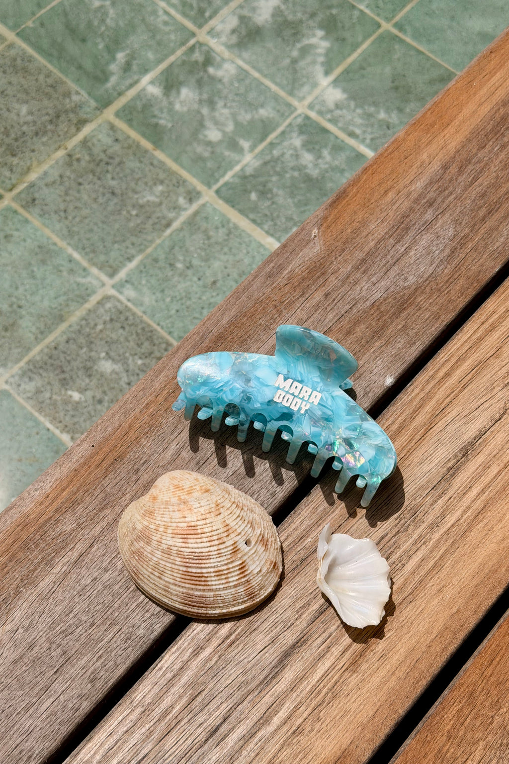 A light blue tortoiseshell hair claw clip with 'Mara Body' printed in white, resting on a wooden deck next to two seashells.