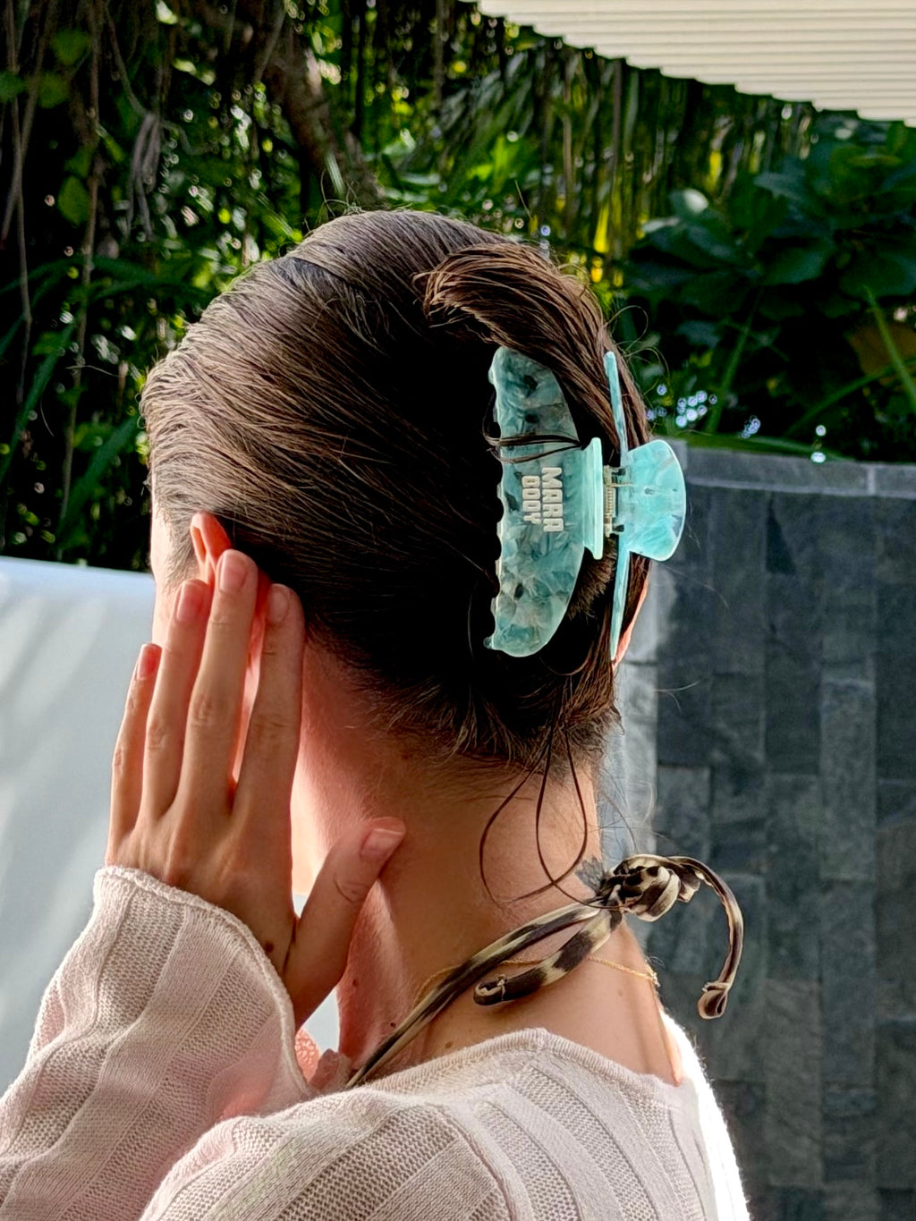 A person shown from the back wearing a large, scalloped-edge claw hair clip in a light teal green color, holding their hair in an updo. The background features lush green foliage and a gray stone wall.