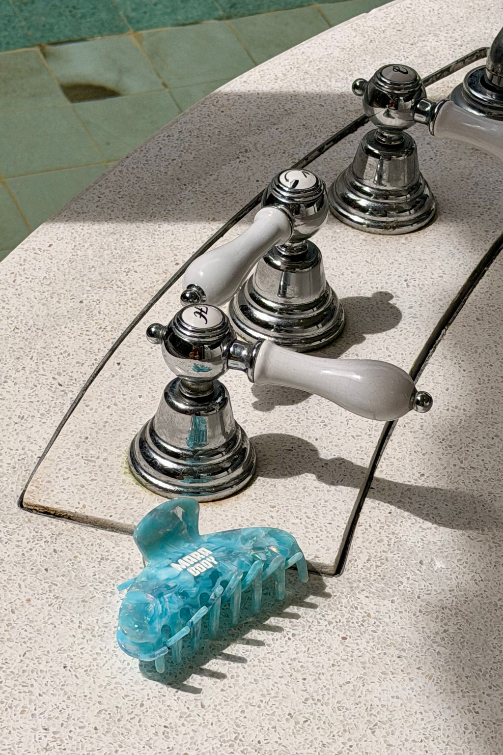 A close-up of a bathroom faucet with polished chrome handles and white lever knobs, mounted on a speckled light beige stone sink. A translucent light blue hair claw clip is placed in the foreground.