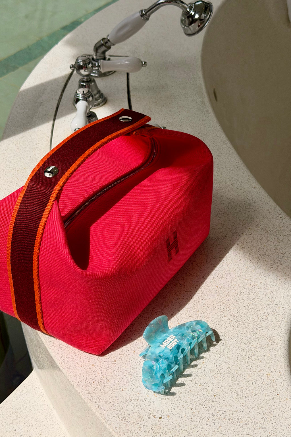 A bright pink red bride a brac with a dark red handle and orange piping sits on a bathroom counter next to a chrome faucet, with a small baby blue hair claw mara body clip in front of it.