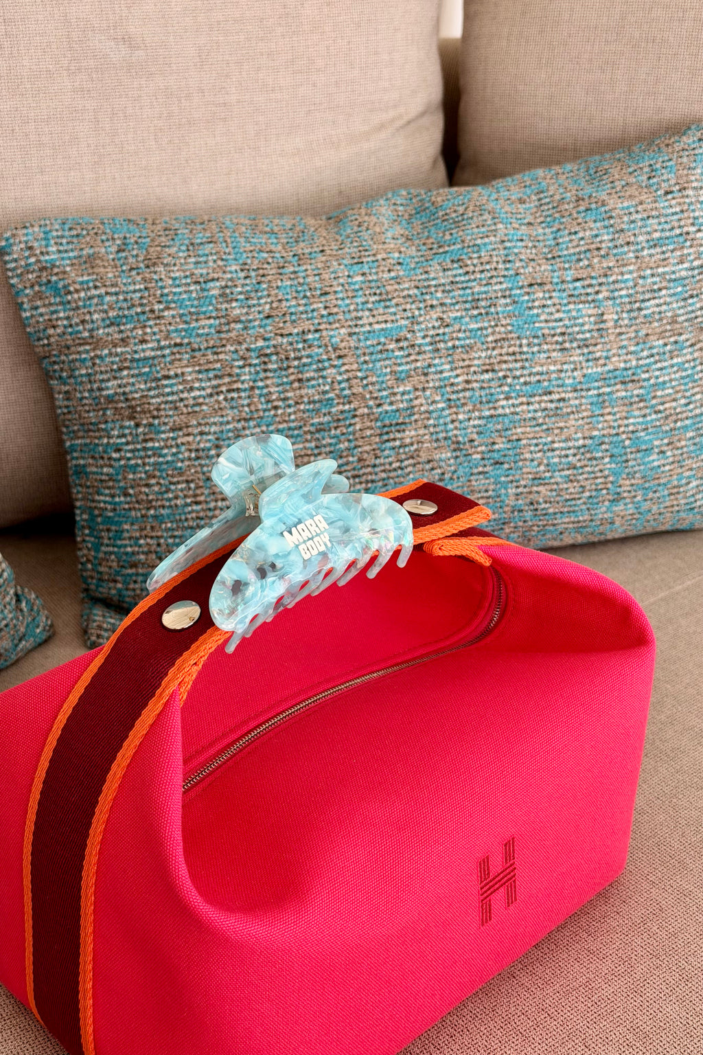 A bright pink cosmetic bag with a dark red handle and orange trim, featuring a light blue tortoiseshell hair claw clip attached to the handle. The bag has a small red embroidered 'H' logo on the front and is resting on a beige sofa with a blue and beige patterned pillow in the background.