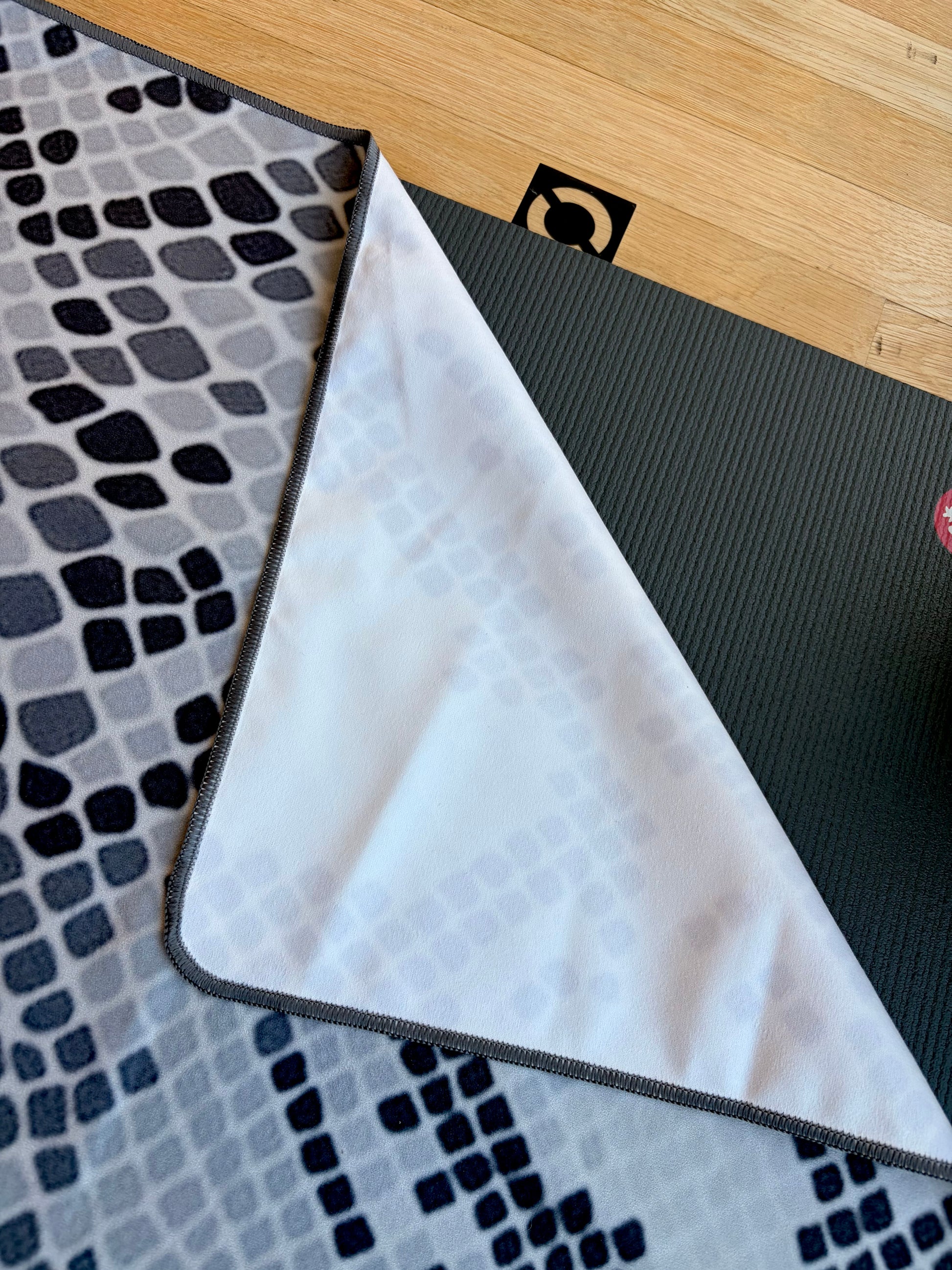 A close-up of a non slip microfibre yoga towel with one corner folded over to show both sides: a dark charcoal ribbed top surface and a white bottom surface with a subtle geometric pattern. The mat is placed on a light wood floor next to a gray and black patterned rug
