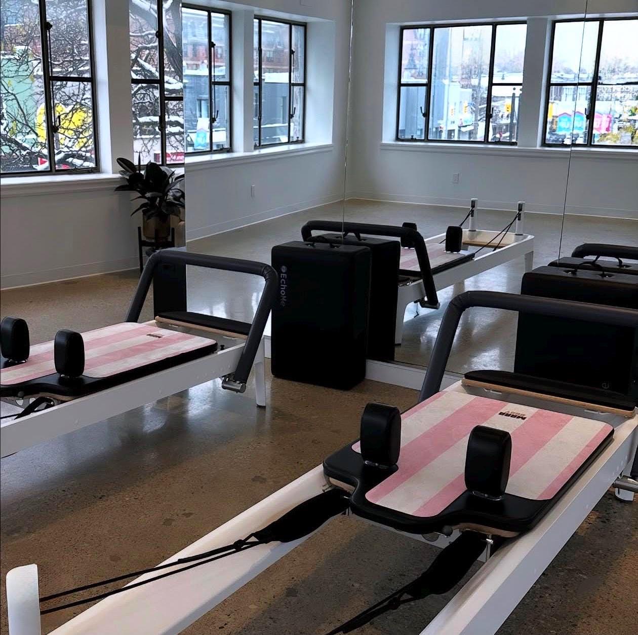 Pilates reformer machines with pink pilates reformer mats arranged in a bright studio with large windows, concrete floors, and mirrored walls.