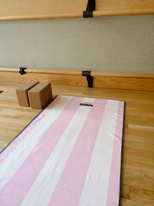 A pink and white striped yoga mat towel with a black border lies on a wooden floor in a studio setting, with two brown yoga blocks and wooden barre rails in the background.