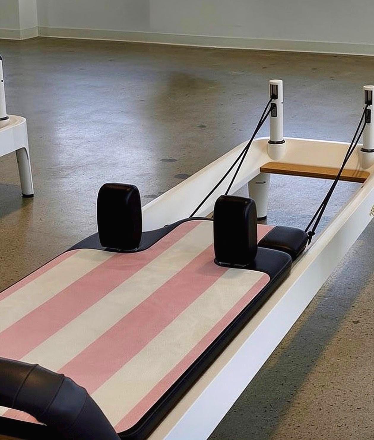 Pilates reformer machine with a pink and cream white striped mat, black handles and foot pad, and a wooden slide, photographed in a studio with a concrete floor.