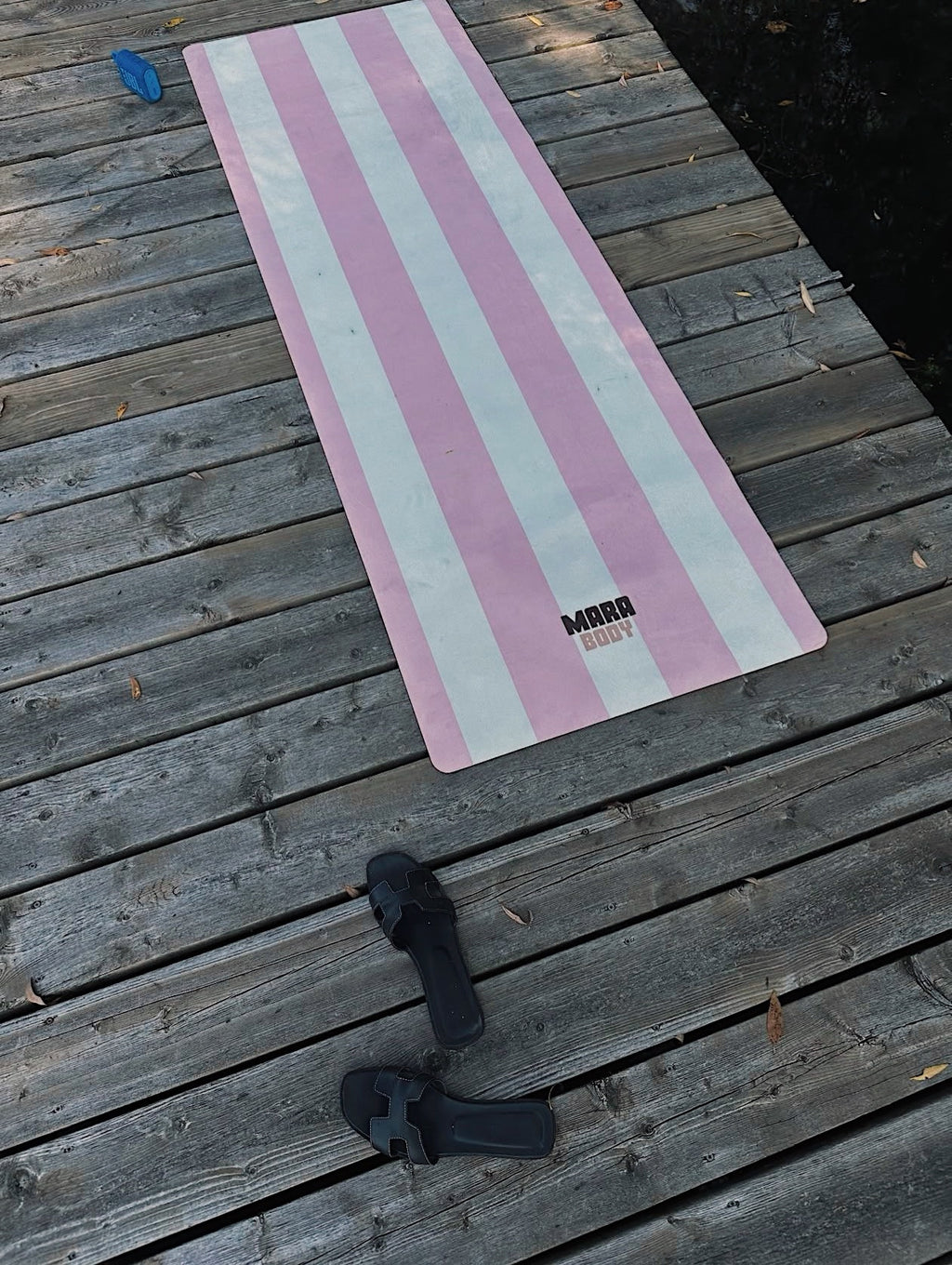 A pink and white striped yoga mat with the brand name 'MARA BODY' printed near the bottom, laid out on a weathered wooden deck. A pair of black sandals hermes and a blue cap are placed nearby on the deck.