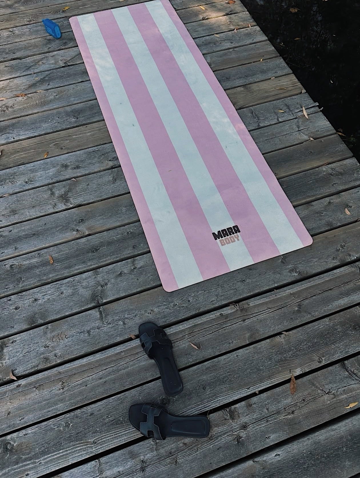 A pink and white striped yoga mat with the brand name 'MARA BODY' printed near the bottom, laid out on a weathered wooden deck. A pair of black sandals hermes and a blue cap are placed nearby on the deck.