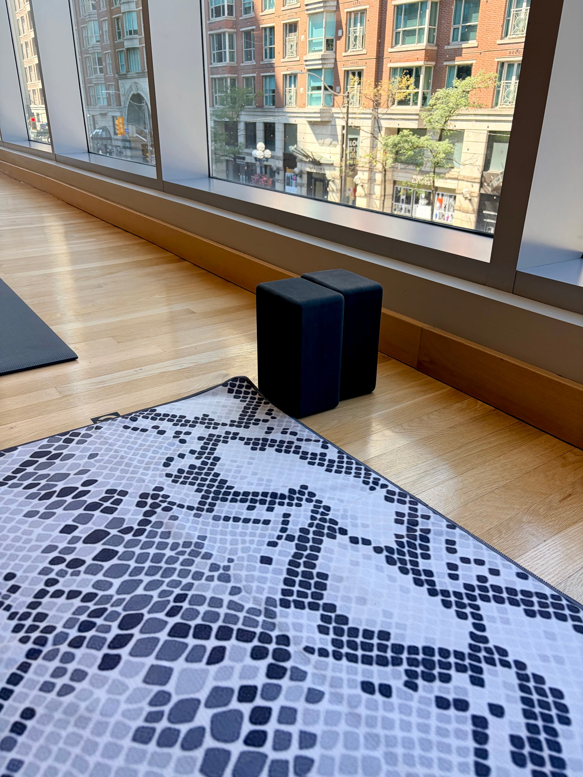 A black and white snakeskin-patterned yoga towel for yoga mat on a light wood floor in a bright room with large windows and two black yoga blocks.
